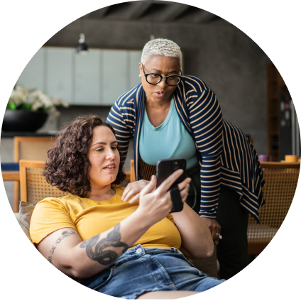 A white woman lays on a couch holding up her phone, while her partner, a Black woman in glasses, leans over behind her, peering at the same phone.