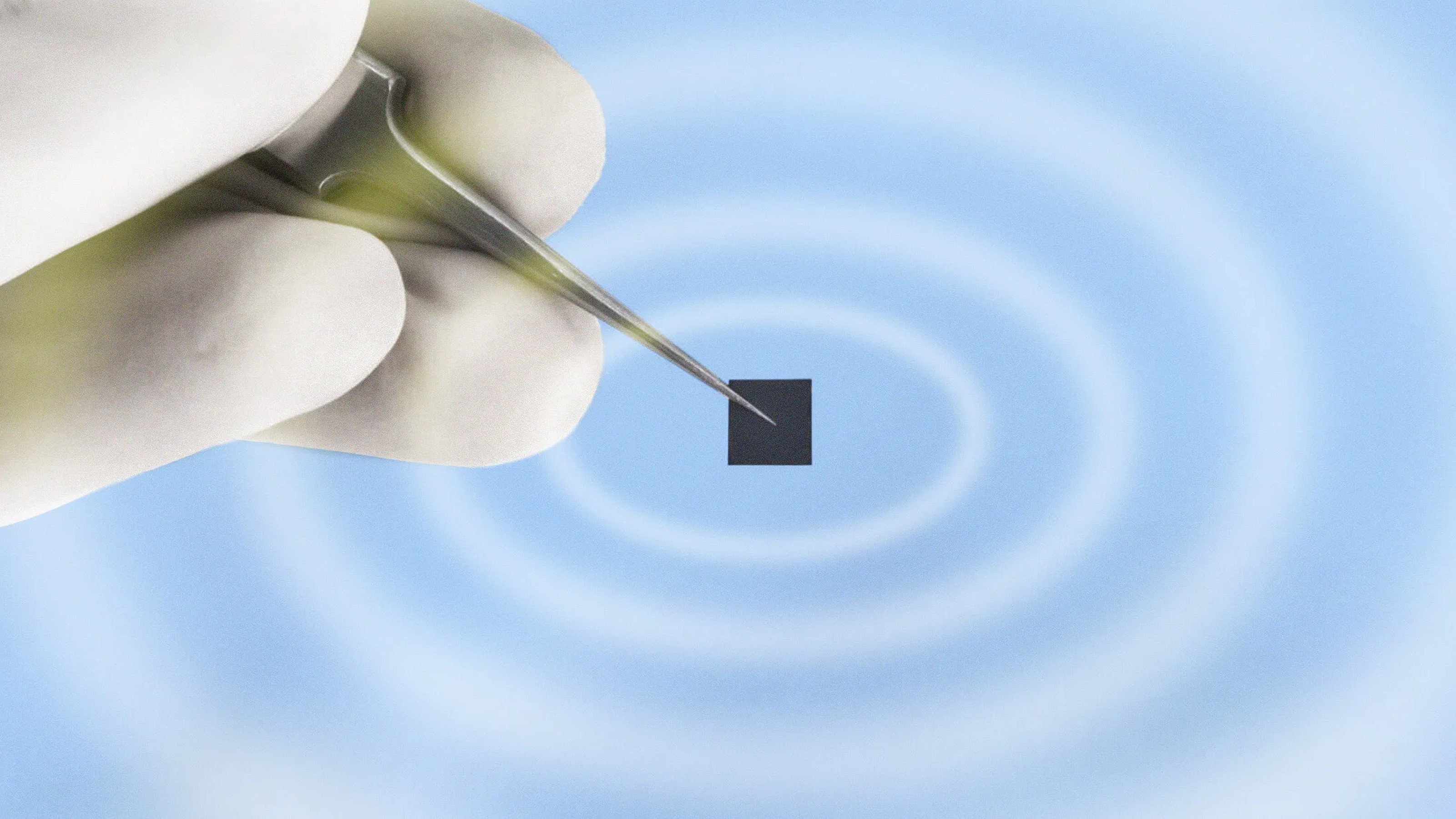 A hand holding tweezers grips a small black square against a blue background with concentric white circles.