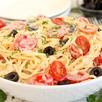 White serving bowl filled with creamy angel hair pasta with tomatoes and black olives.