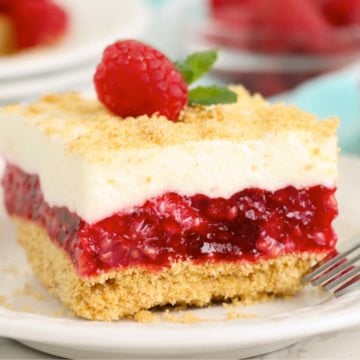 Piece of raspberry cake garnished with a fresh raspberry and mint leaf on top of a white plate.