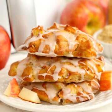 Three apple fritters stacked on a white dessert plate in front of an air fryer.