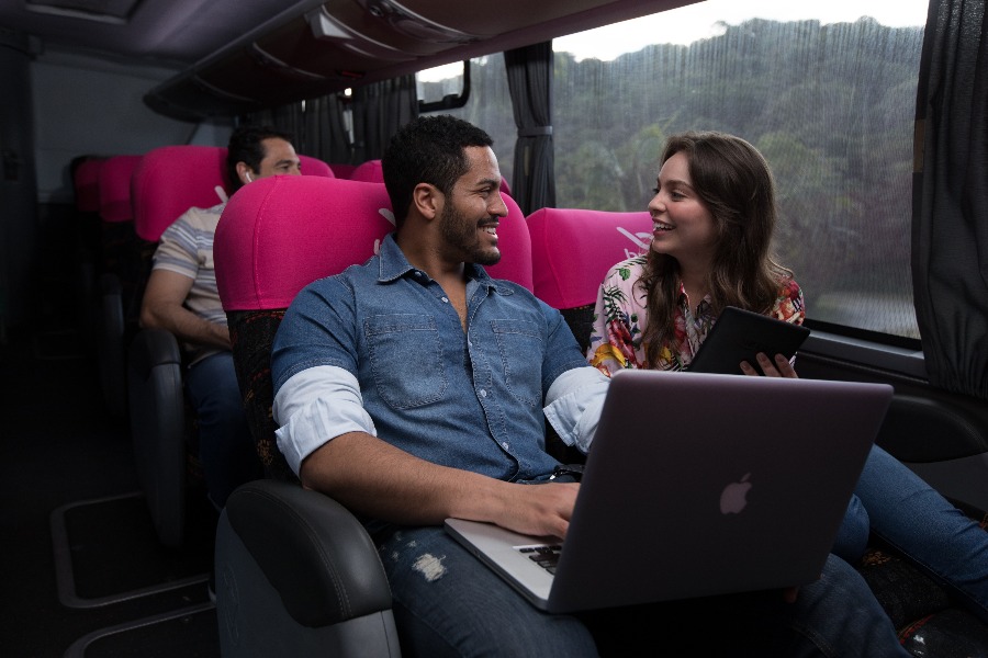 poltrona de ônibus leito-cama. Na foto, home negro com um notebook viajando ao lado de uma mulher morena de cabelos lisos.