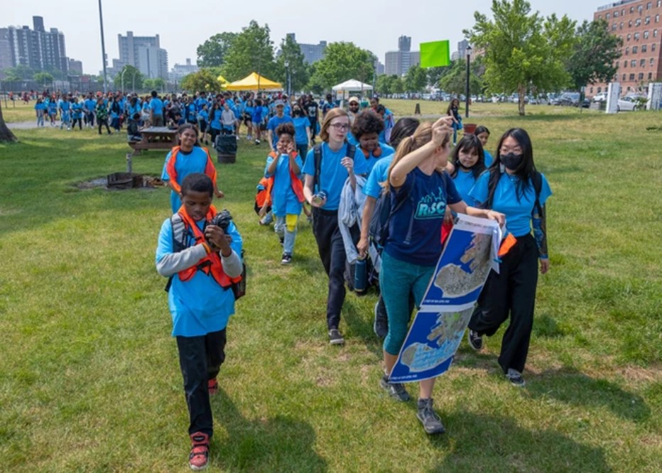 A group of young people march over a grass field holding posters and banners. They are all wearing blue t-shirts.