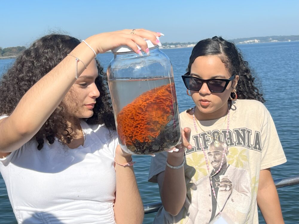 Two students on a boat look at a specimen in a large jar filled with water. One student is wearing sunglasses.