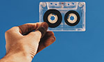 Photograph of a hand holding a cassette tape against a blue background.