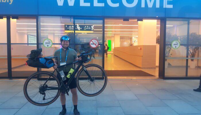 A happy ben lifting up his bike in front of the conference center under a sign saying 'Welcome'