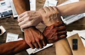 People of different races holding hands over a desk, depicting a diverse workforce.