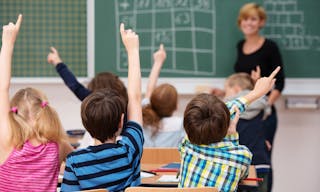 Intelligent group of young school children all raising their hands in the air to answer a question posed by the female teacher, 