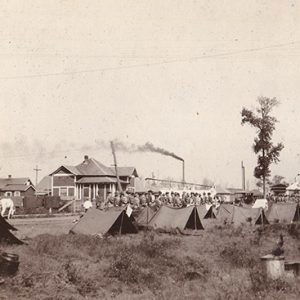 Men standing near rows of tents and water tower with town buildings in background