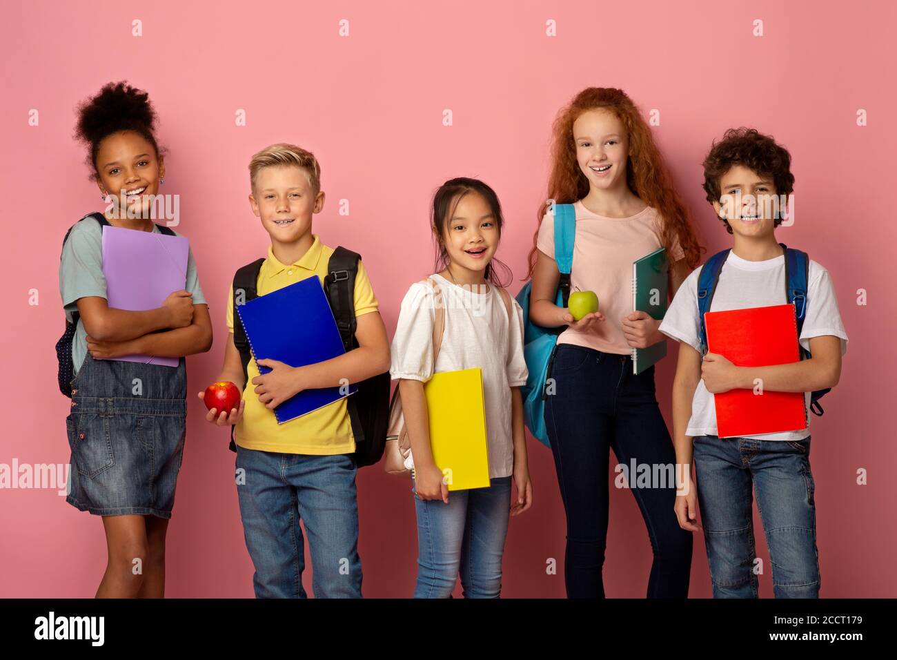 Getting ready for new school year. Diverse children with backpacks, notebooks and apple fruits on pink background Stock Photo