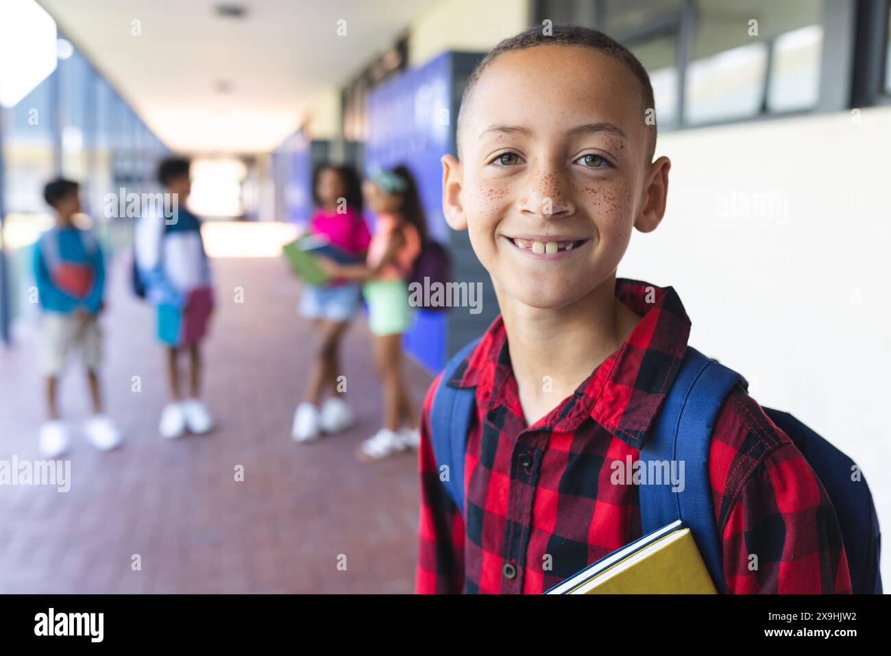 A biracial boy with a red plaid shirt and backpack smiles at school Stock Photo