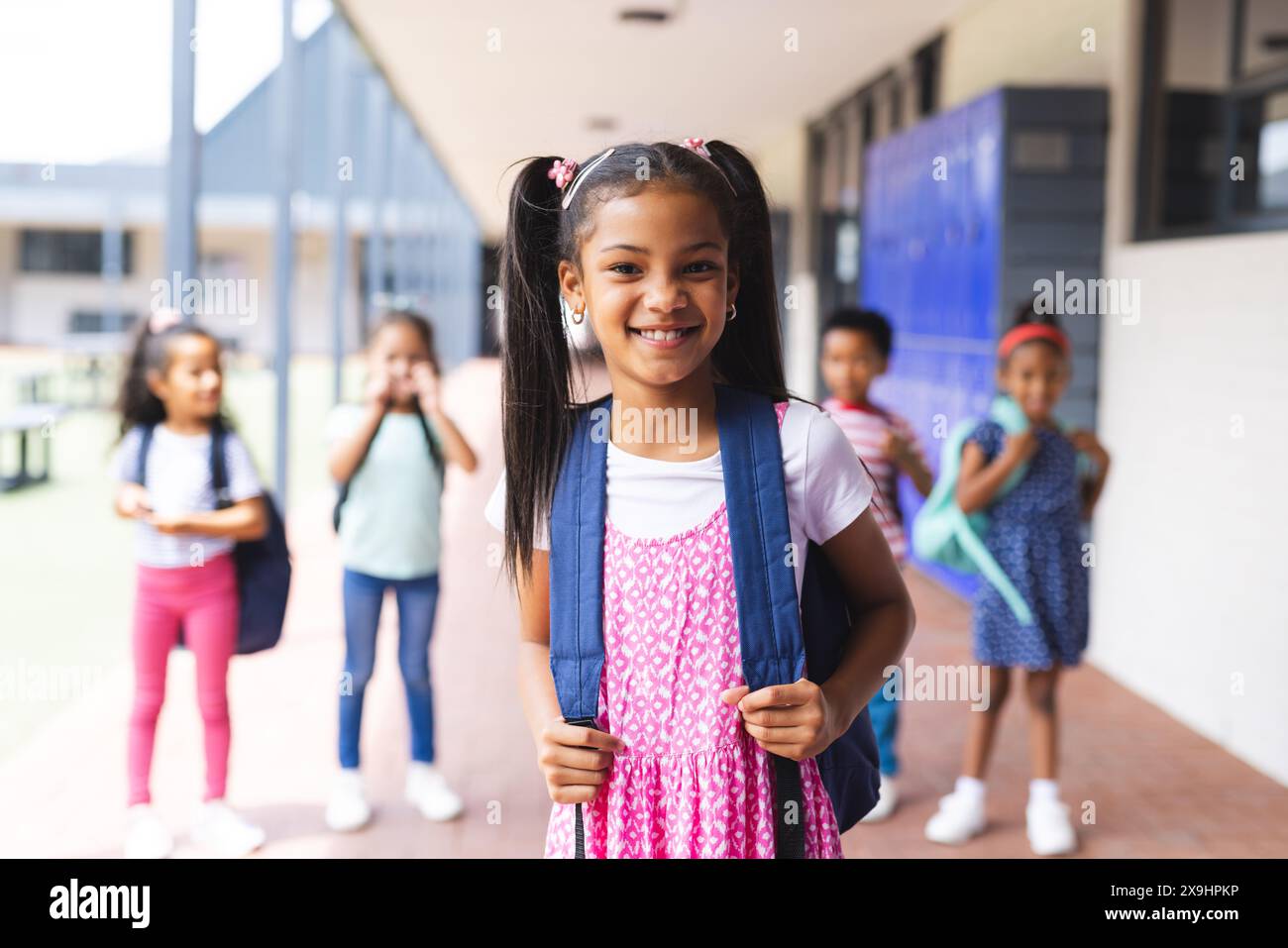 In school, young biracial girl with pink dress and backpack is smiling outdoors Stock Photo