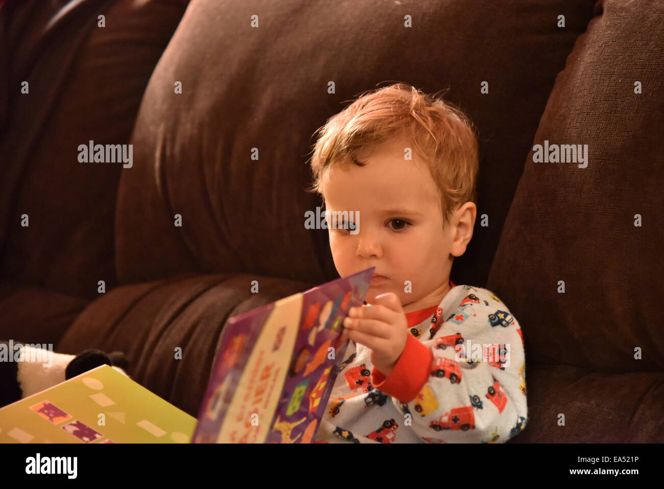 A young boy (two and a half years old) in pyjamas sat on a sofa reading a book just before bedtime Stock Photo