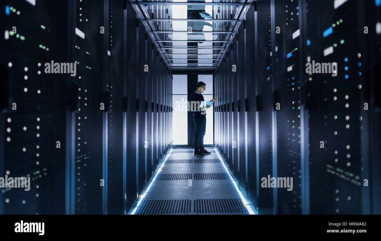 Male IT Engineer Works on a Laptop at the end of a Corridor in a Big Data Center. Rows of Rack Servers are Seen. Stock Photo