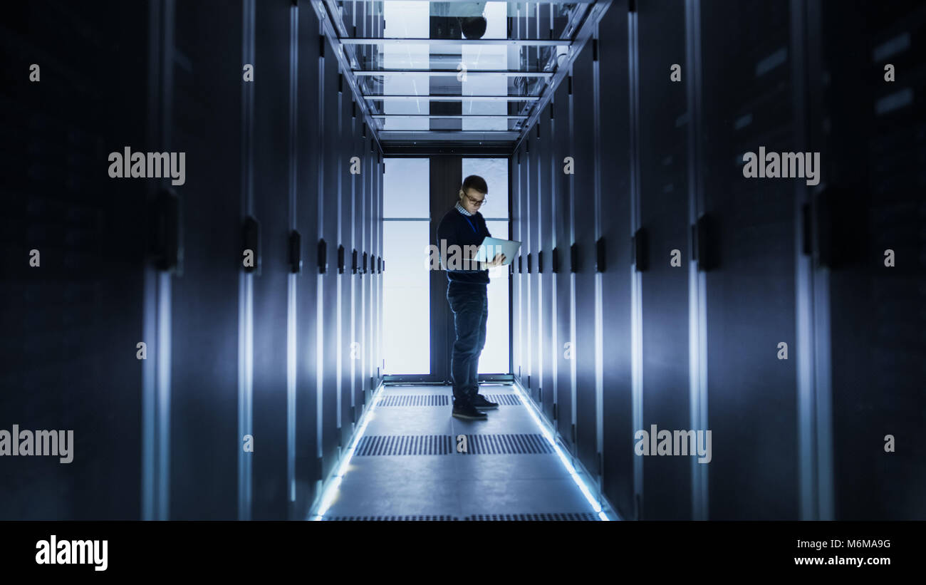 Male IT Engineer Works on a Laptop at the end of a Corridor in a Big Data Center. Rows of Rack Servers are Seen. Stock Photo