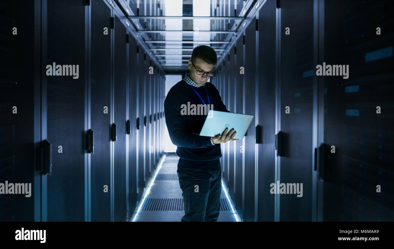 Male IT Engineer Works on a Laptop in front of Server Cabinet at a Big Data Center. Rows of Rack Servers are Seen. Stock Photo