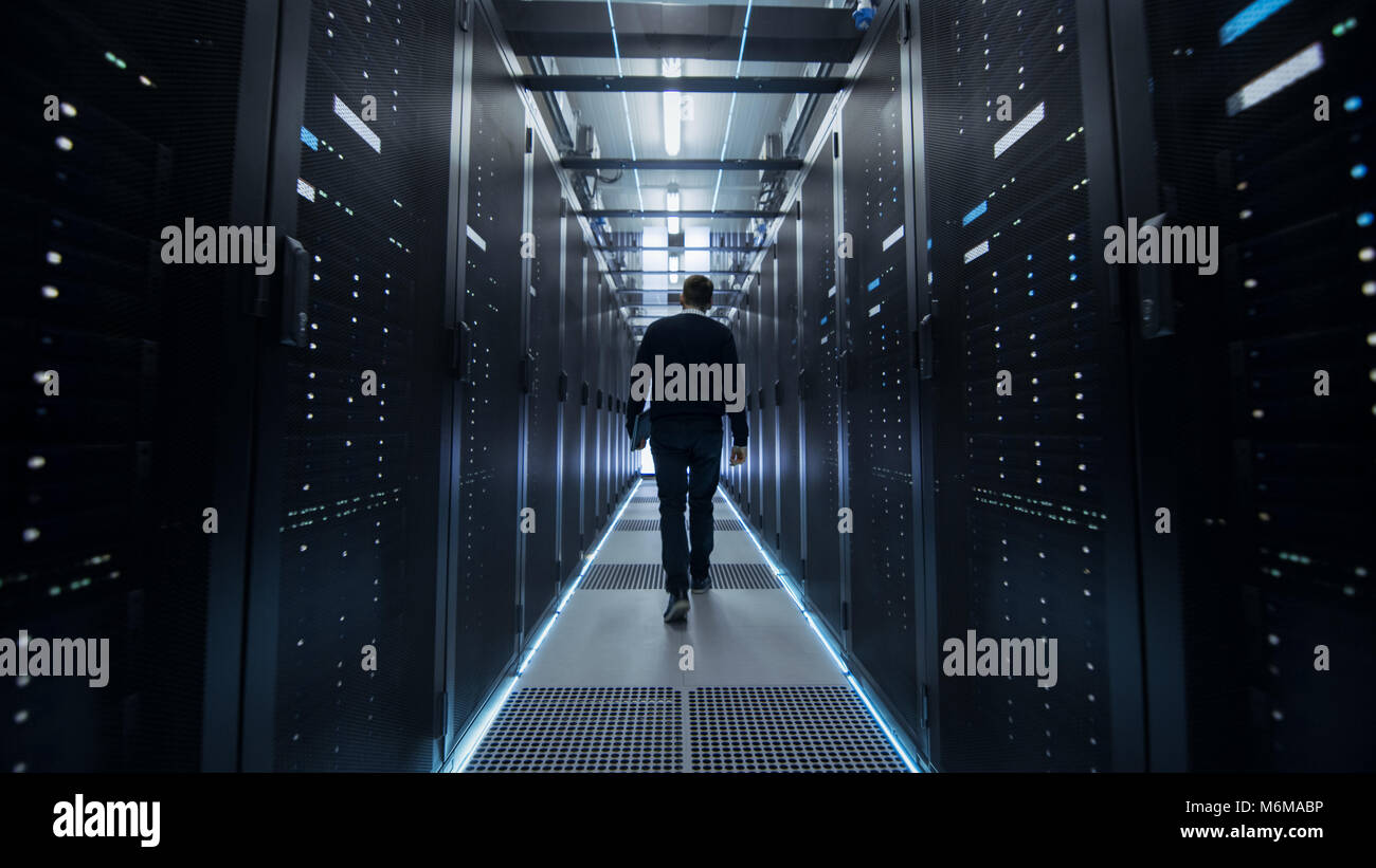 Following Shot of IT Engineer Walking Through Data Center Corridor with Rows of Rack Servers. Stock Photo