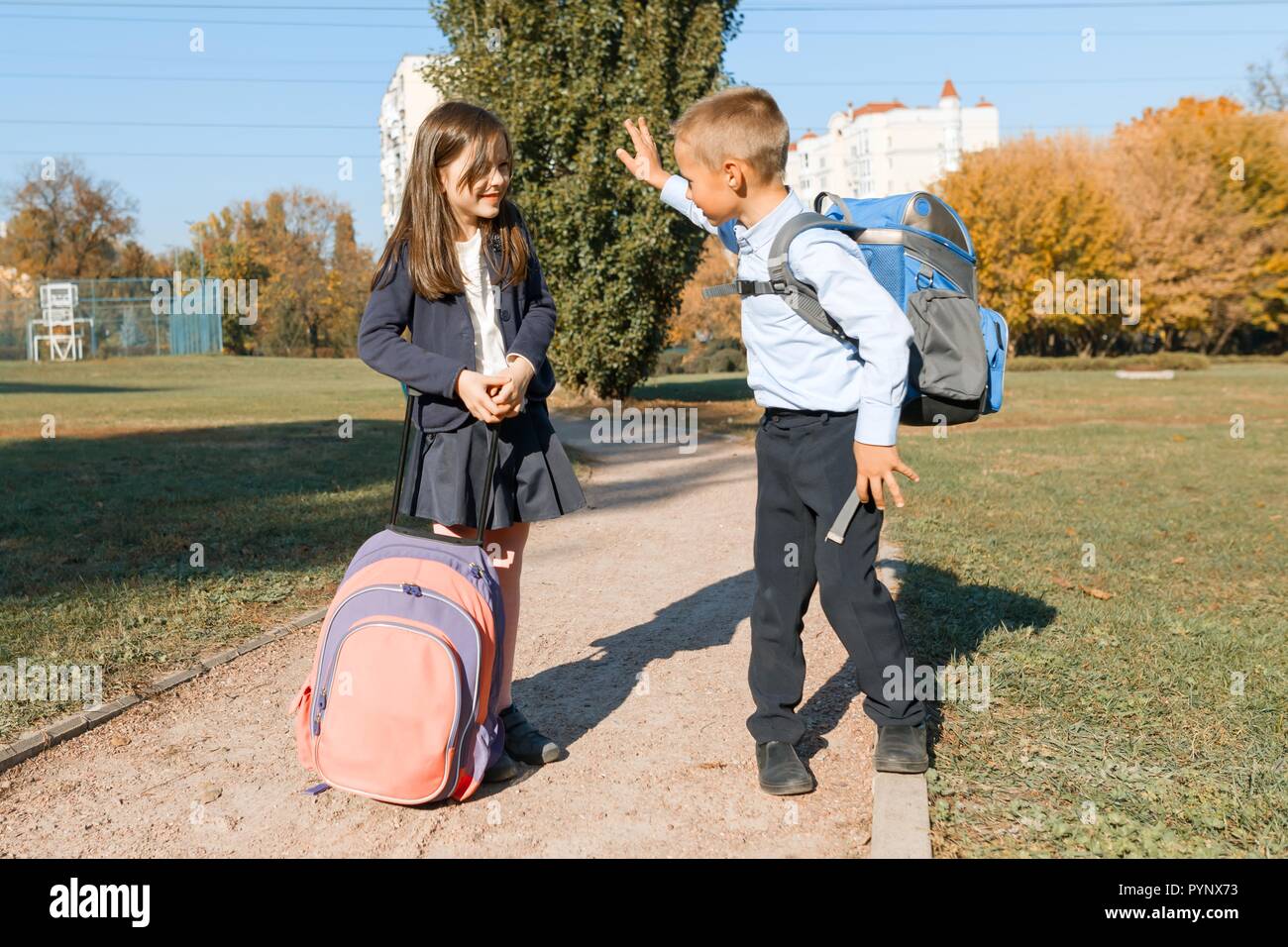 Boy and girl, primary school students with backpacks go to school. Sunny day background, road in the park. Stock Photo