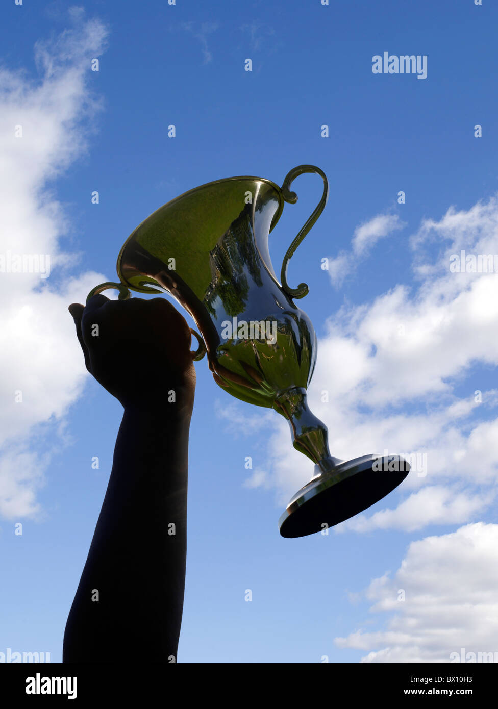 Eine siegreiche Person hält einen großen, glänzenden Trophäenbecher hoch, der sich vor einem leuchtend blauen Himmel mit ein paar Wolken bildet. Stockfoto