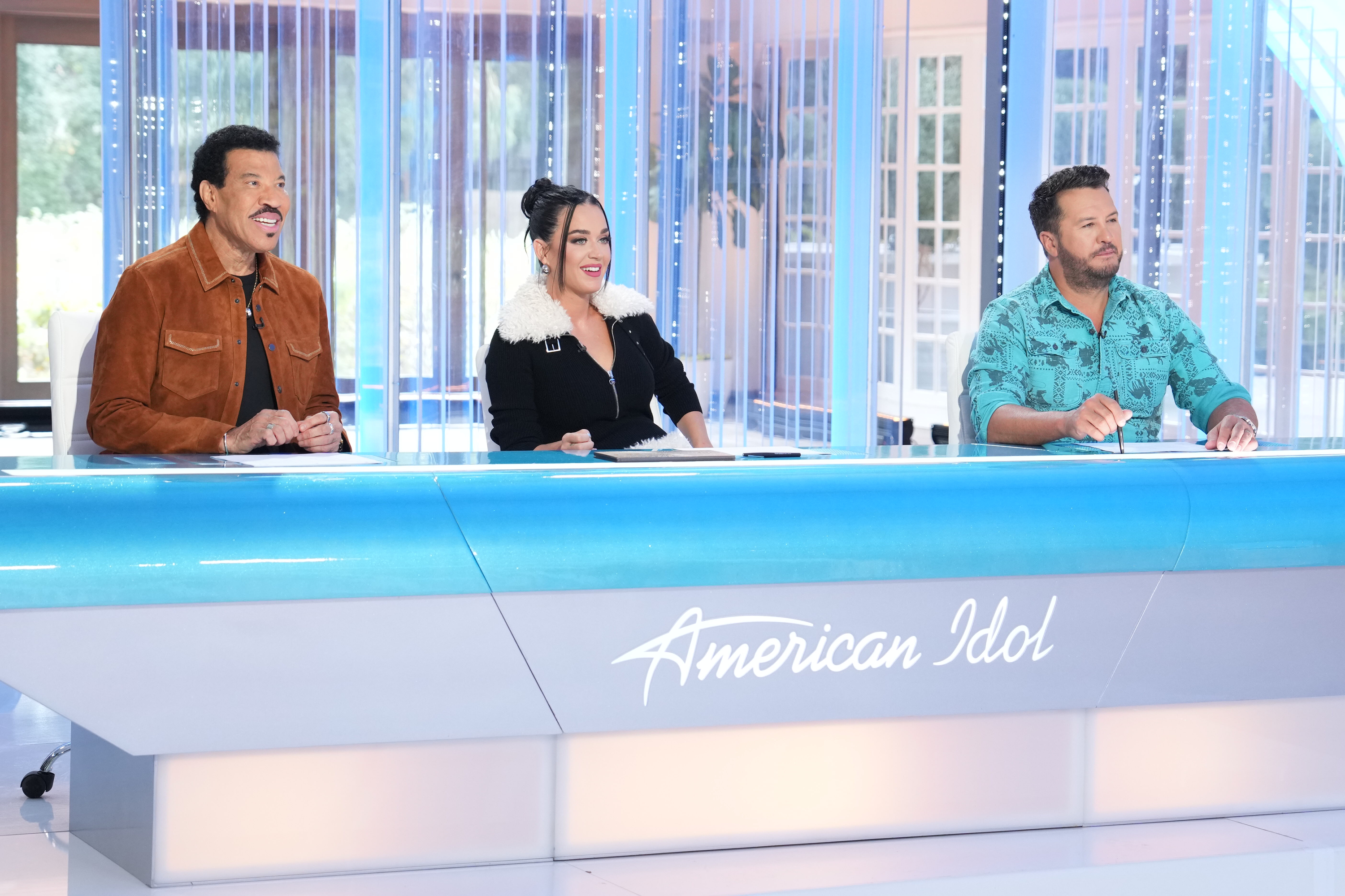 Lionel Richie, Katie Perry and Luke Bryan sitting behind a blue American Idol desk looking ahead of themselves