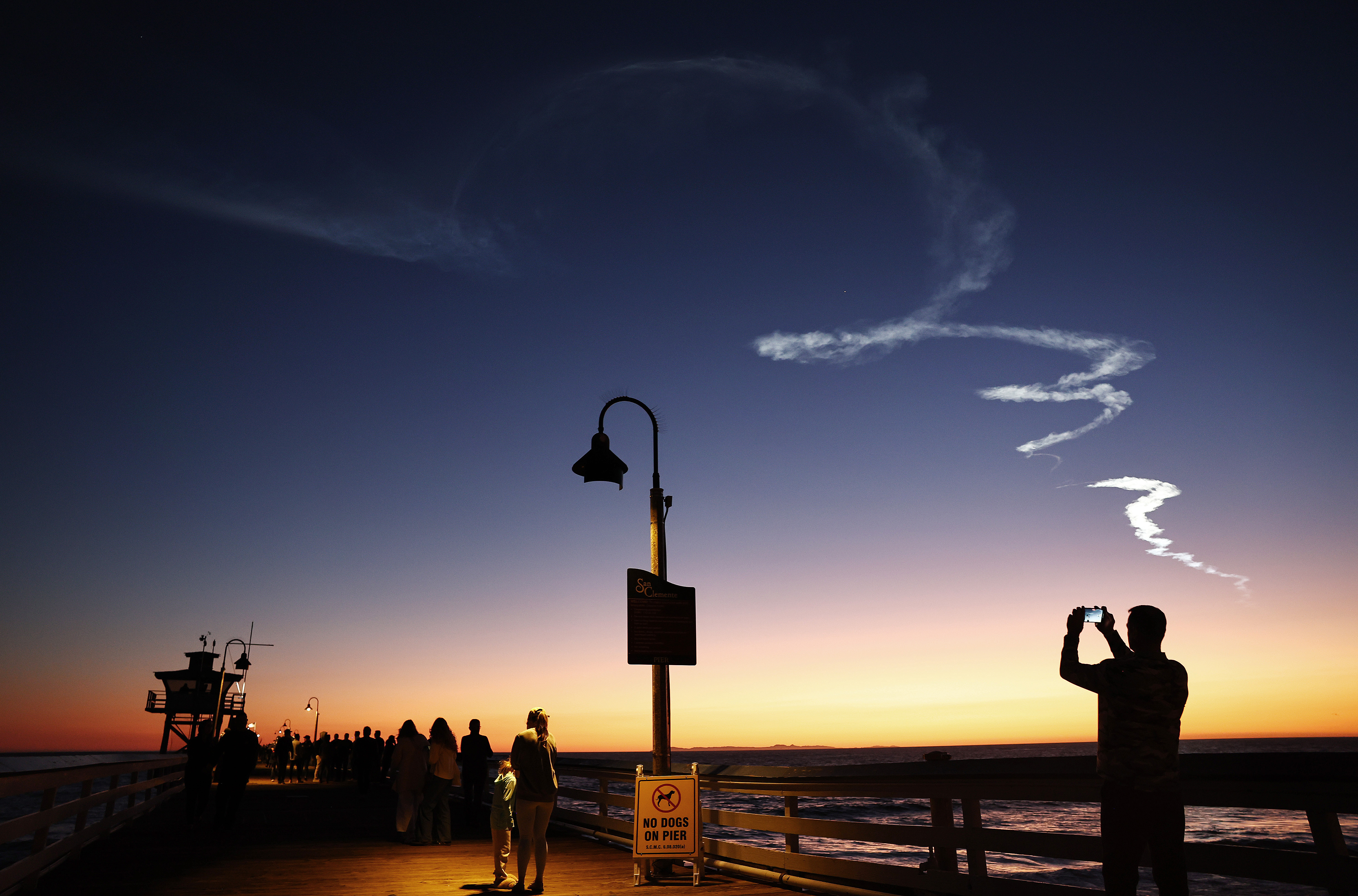 People are silhouetted on a San Clemente pier as they photograph a rocket's contrails at dusk