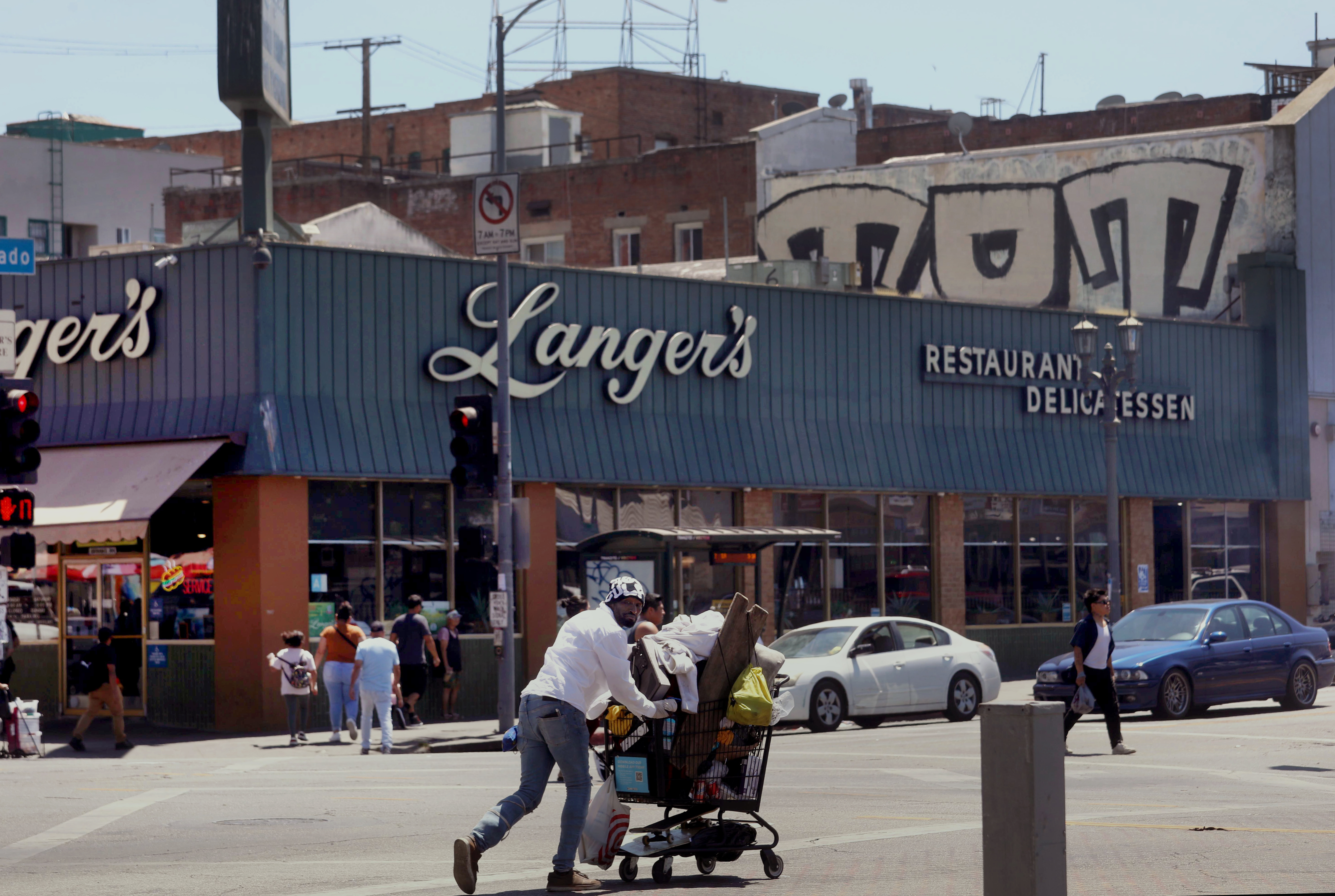 LOS ANGELES, CA - AUGUST 21, 2024 - A man walks with his belongings in a shopping cart across the street from Langer's Deli, also known as Langer's Delicatessen-Restaurant, in in the Westlake neighborhood of Los Angeles, opposite MacArthur Park, on August 21, 2024. Norm Langer, 79, president and chief executive of Langer's Delicatessen-Restaurant, is thinking of closing the deli after 77 years in business, due to neighborhood challenges. (Genaro Molina/Los Angeles Times)