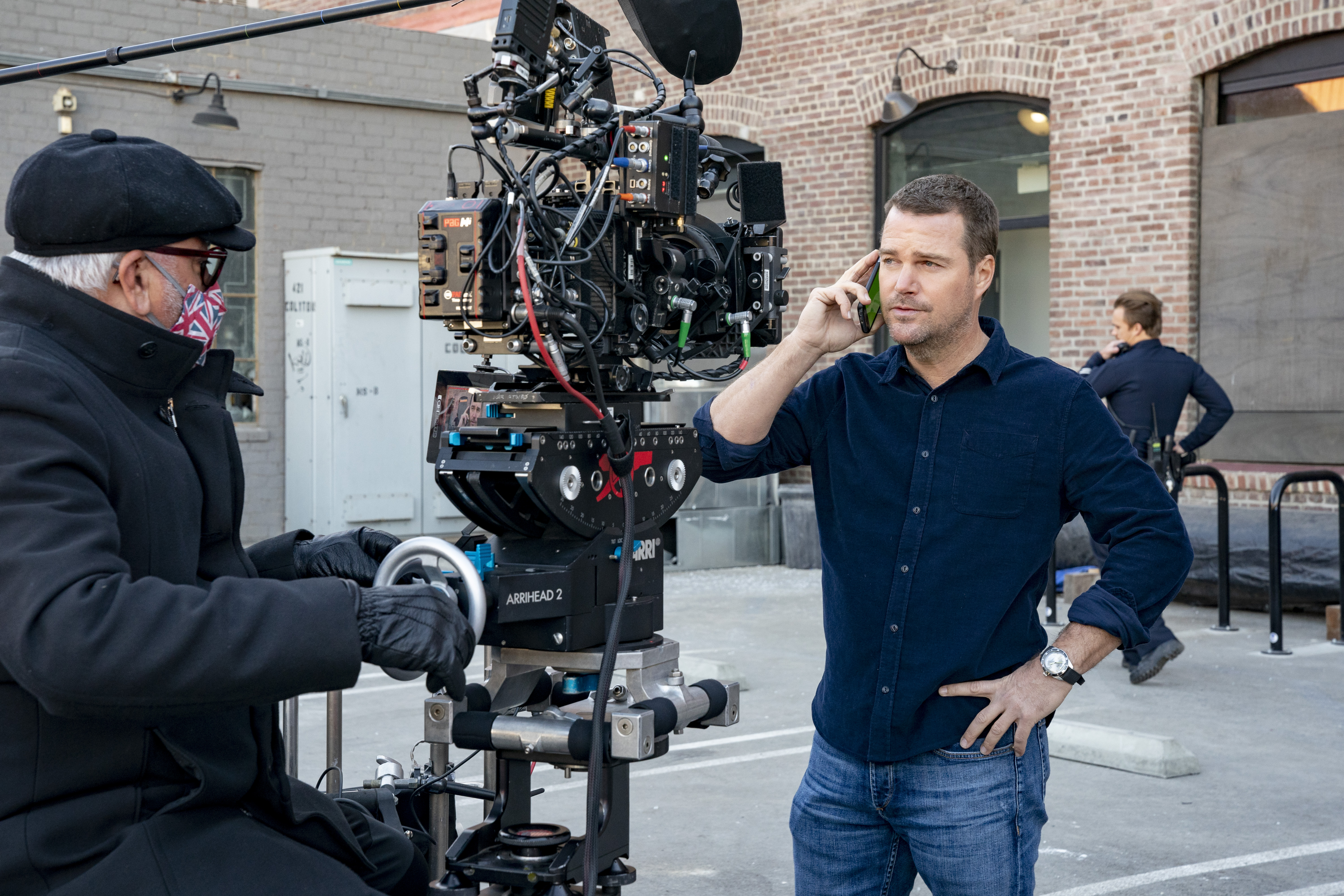 A man wearing a jacket, face mask and a hat filming another man in a collared shirt holding a phone to his ear on a film set