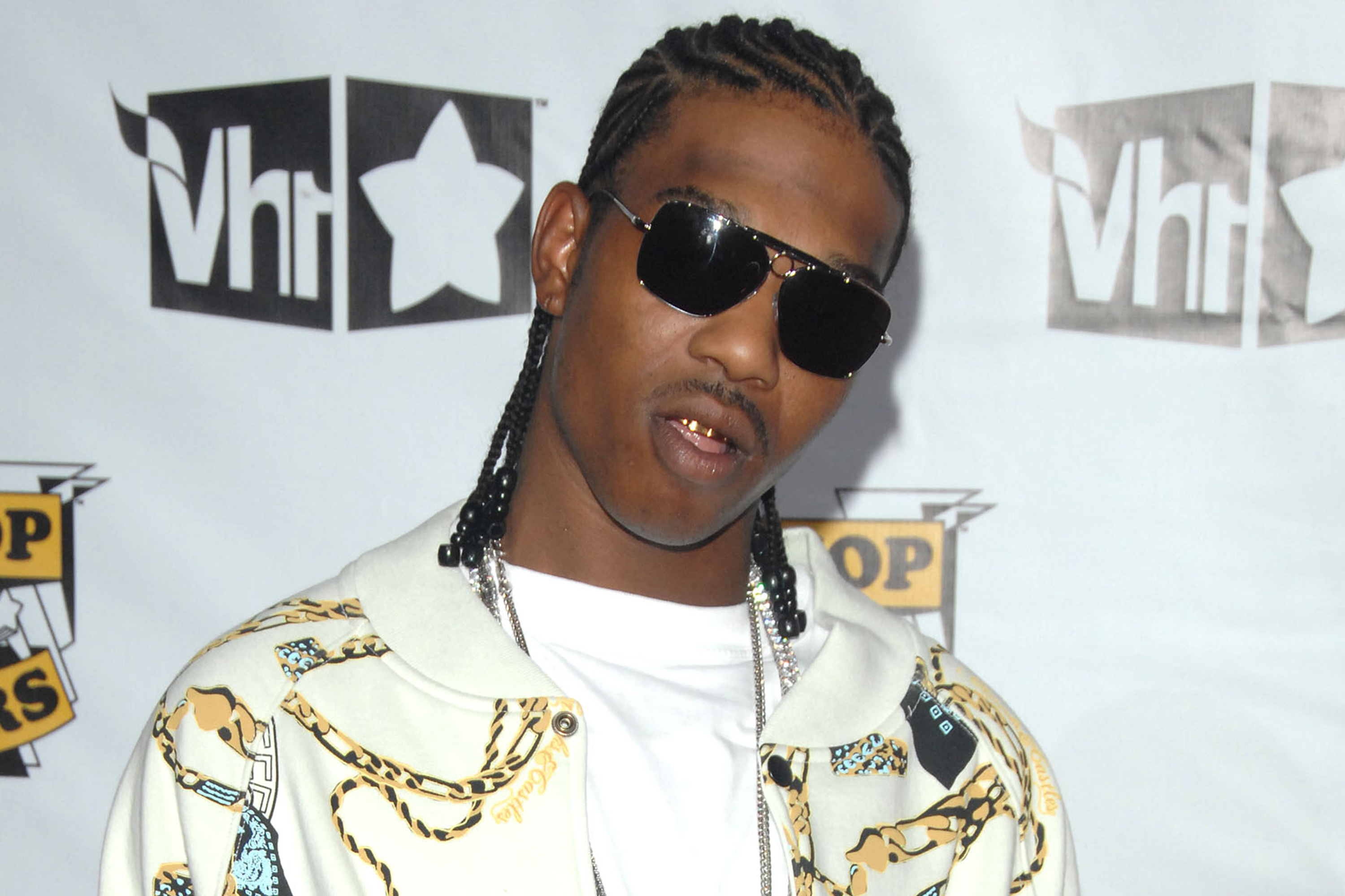 A man wearing cornrows, dark sunglasses, a gold grill, white T-shirt and cream jacket with yellow and blue designs