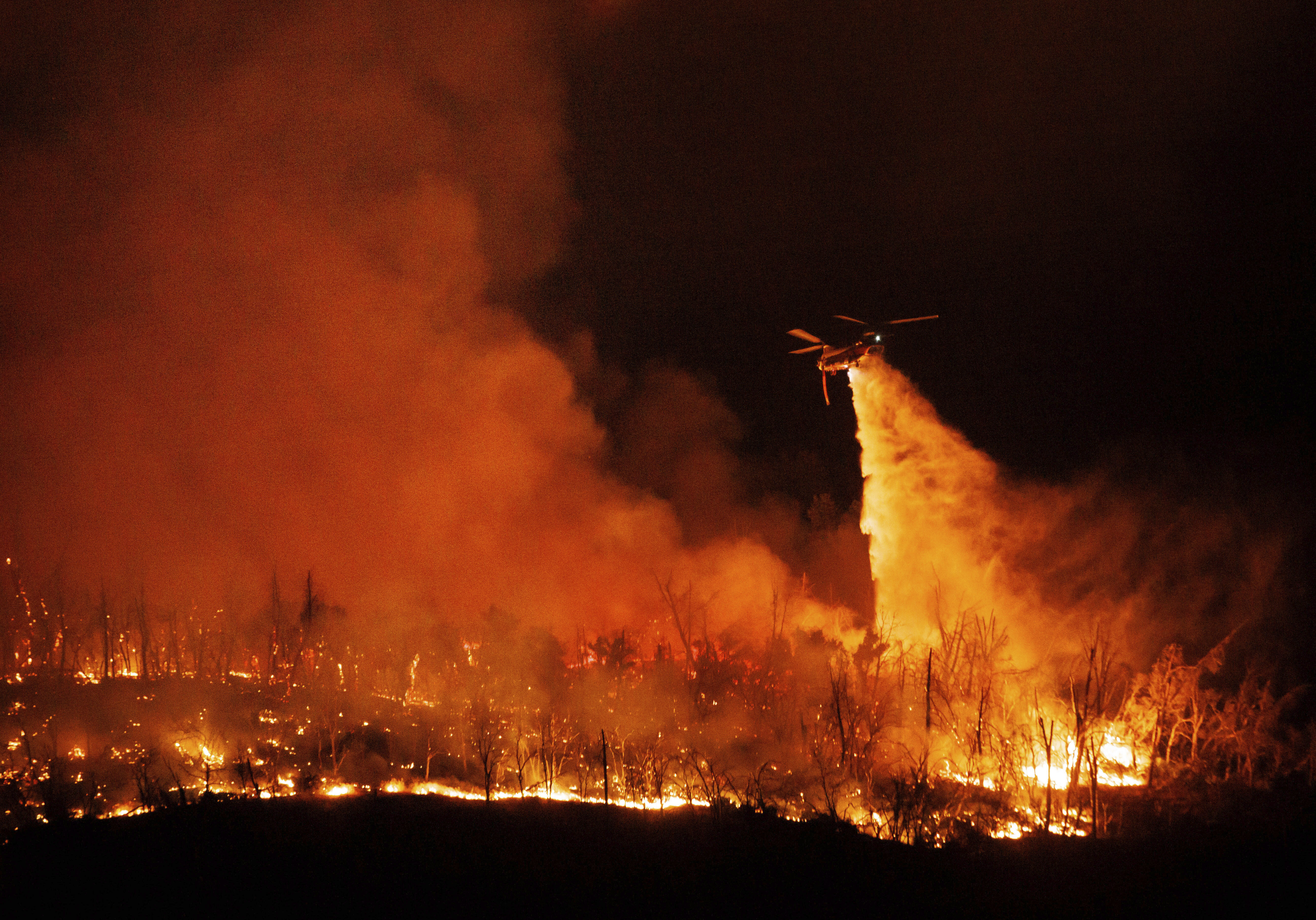 A night flying helicopter drops water on flames as the Thompson Fire burns, Tuesday, July 2, 2024, in Oroville, Calif. (AP Photo/Ethan Swope)