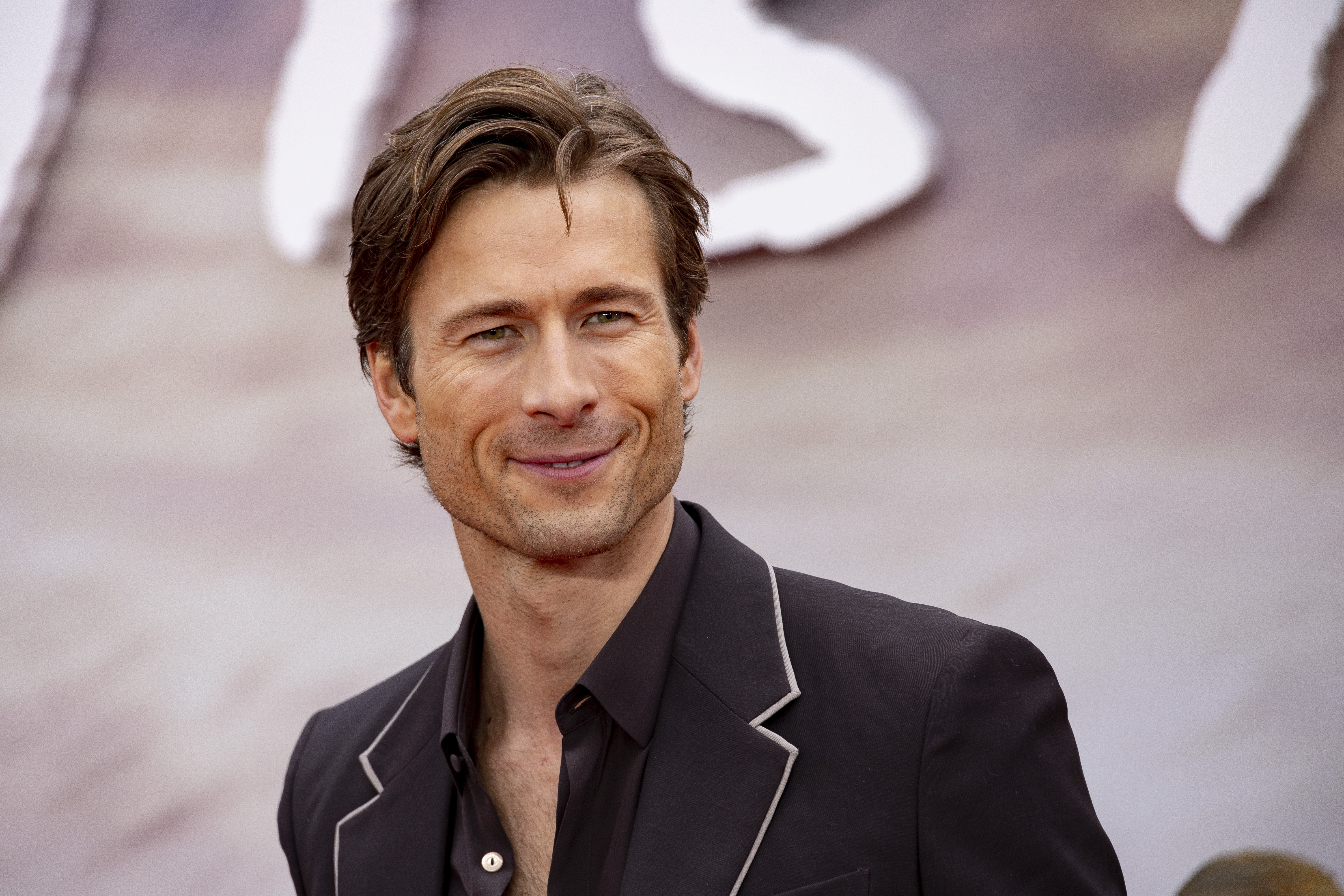 Glen Powell smiles in a black suit at the London premiere of "Twisters" on Monday, July 8, 2024