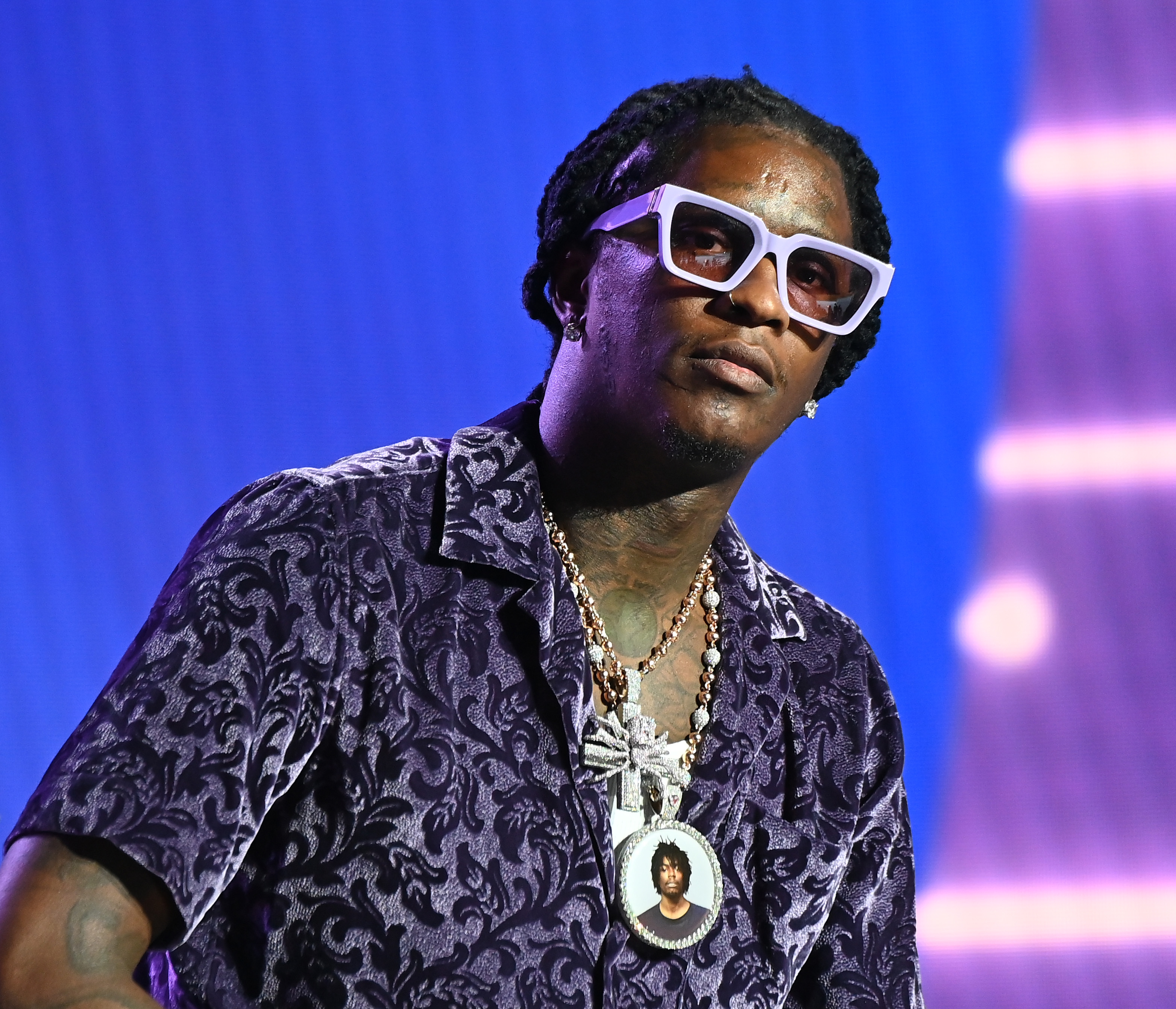 A Black man with thick, white glasses, a dark patterned shirts and chains leaning forward and looking ahead