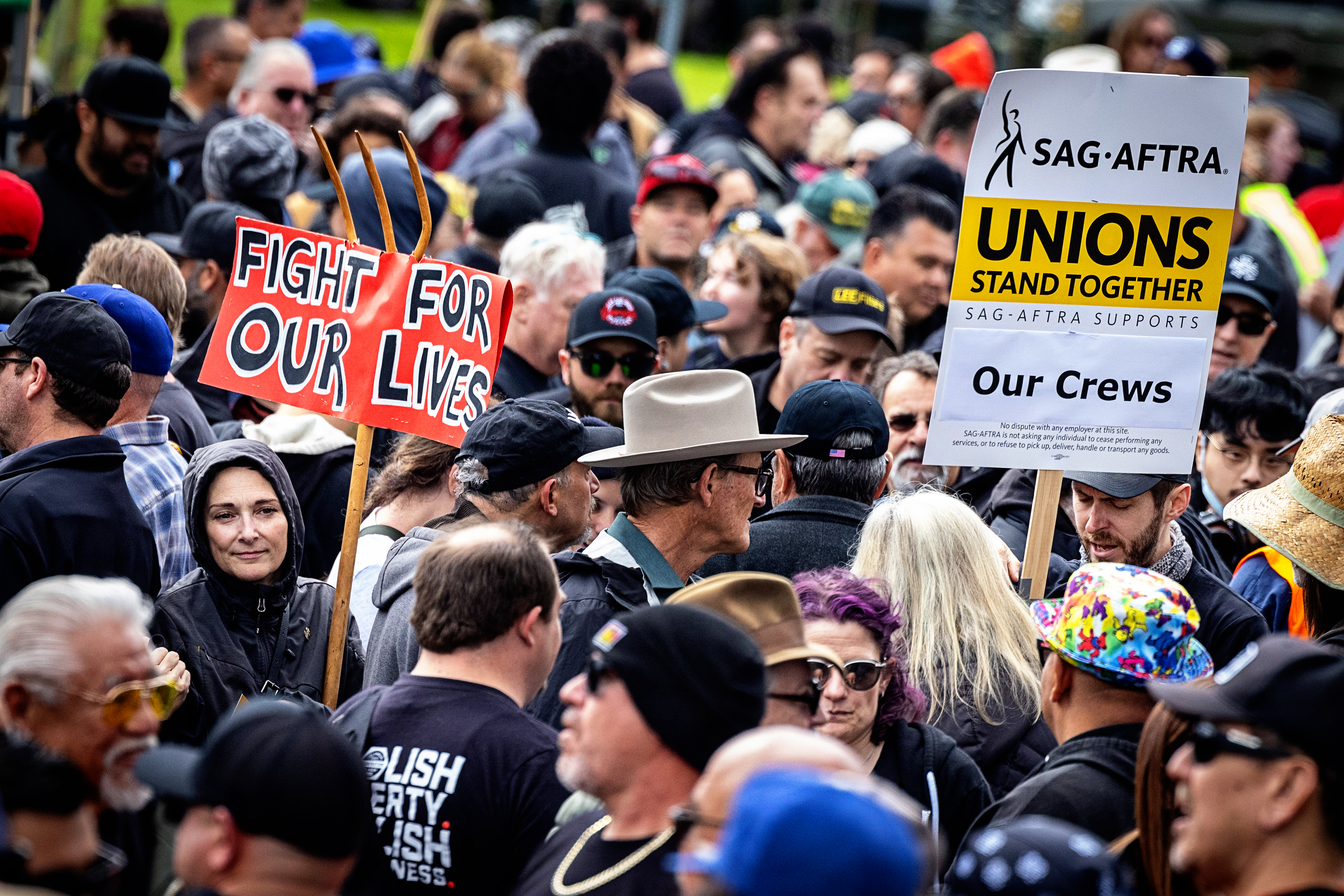 A crowd of people, including two carrying picket signs that read 'Fight for our lives' and 'SAG-AFTRA ... supports our crews'