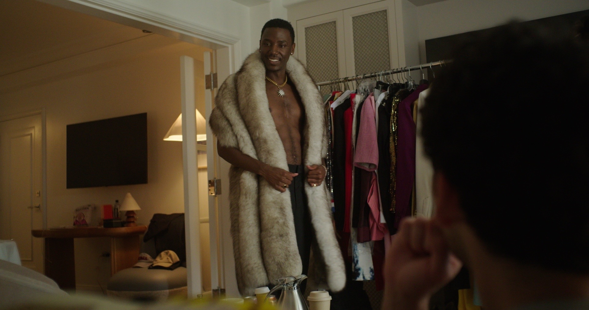 A shirtless man in a fur coat standing before a rack of clothes.