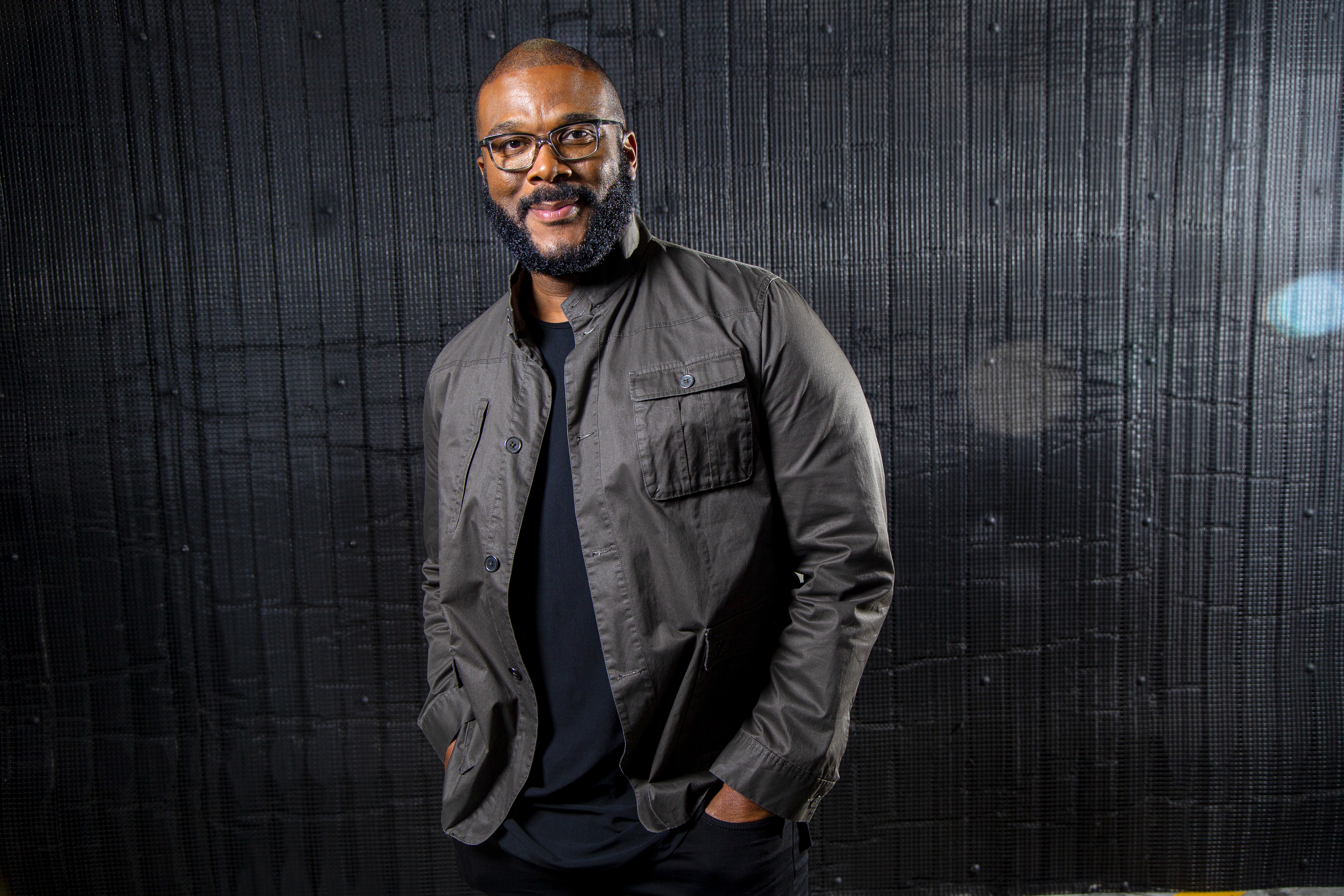 Tyler Perry smiling in a slate gray jacket with his hands in his pockets in front of a black background