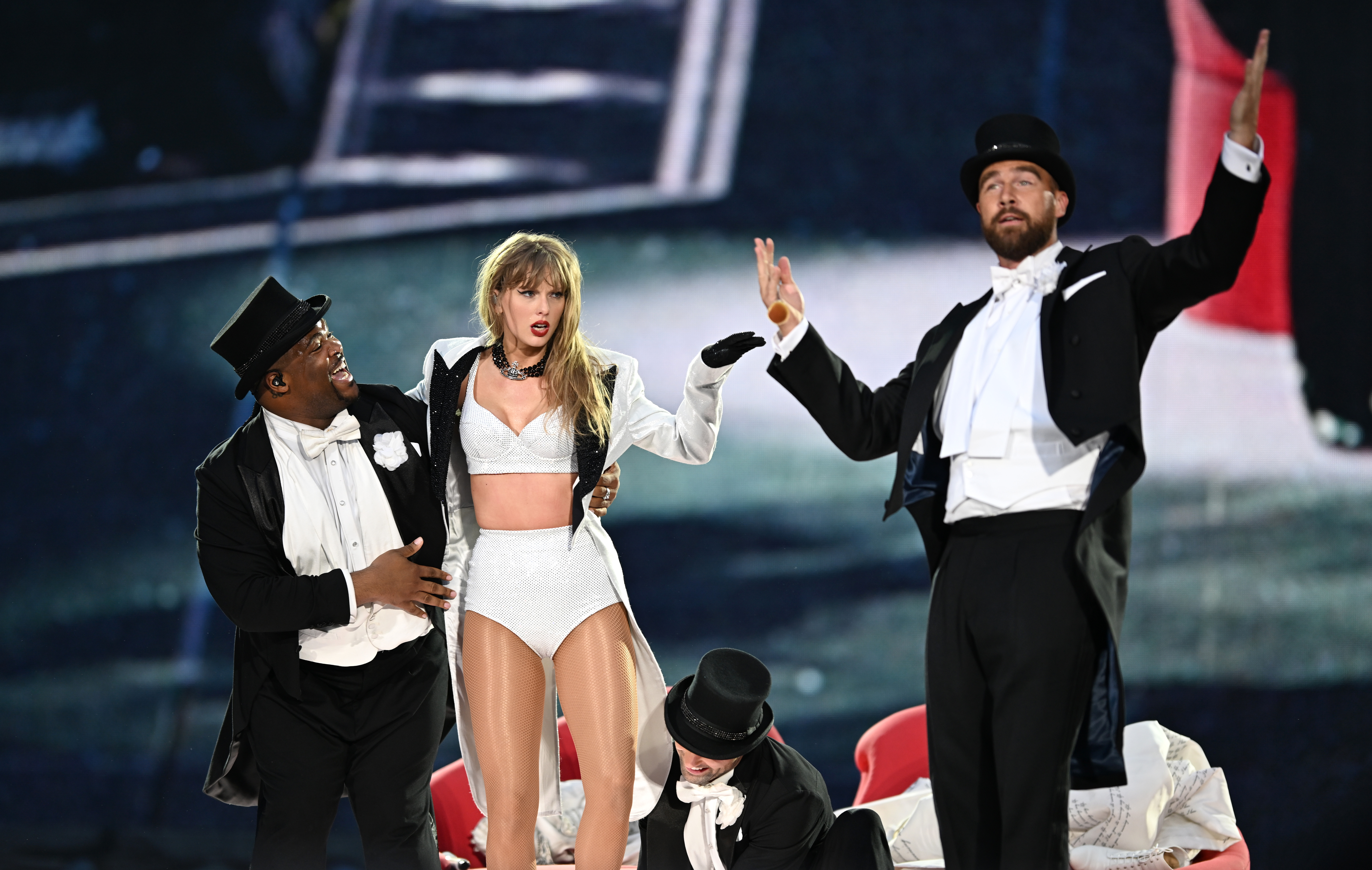 Taylor Swift performing onstage and is joined by Travis Kelce in a top hat and tuxedo