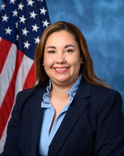 Headshot of Dr. Yadira Caraveo in a dark blue blazer and a light blue blouse with an American flag and blue background.