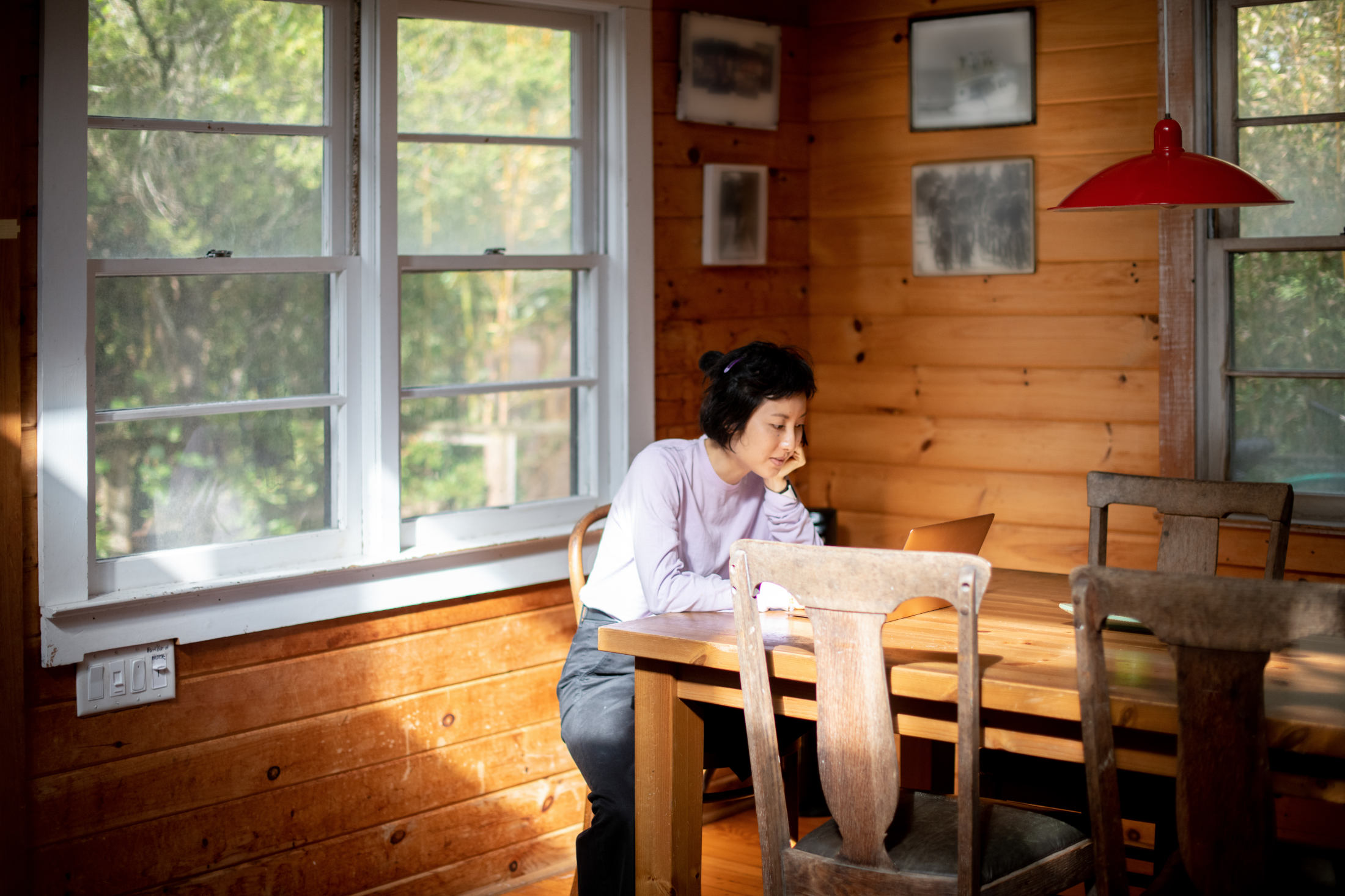 Remote team member working on a laptop at the end of a long wooden table. The room has wooded walls and is naturally lit from the windows