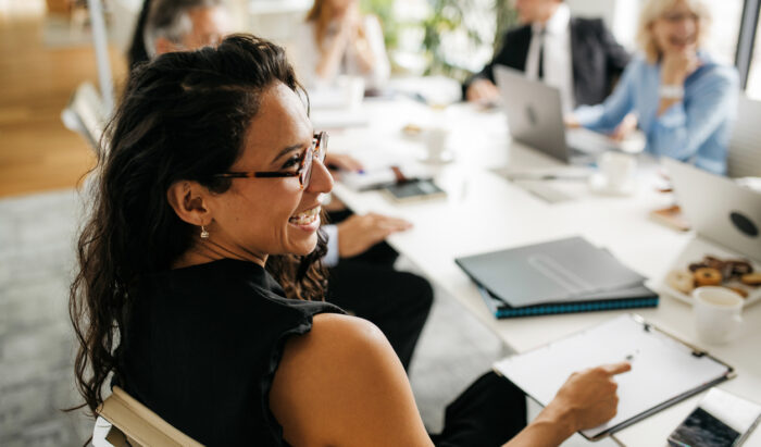 Over the shoulder profile of bespectacled female executive in early 30s sitting at conference table and laughing as she interacts with off-camera colleague.