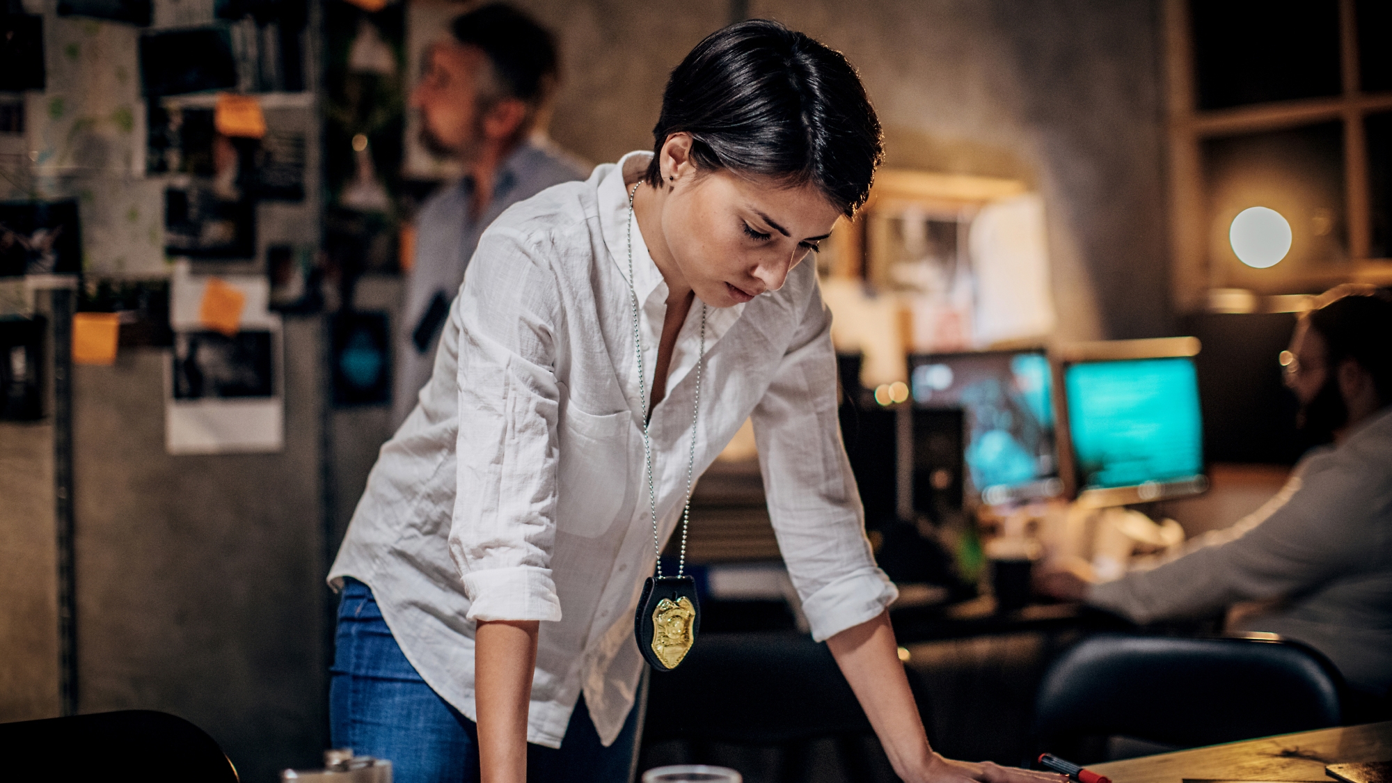 A focused female detective with a badge leans over a desk in a dimly lit office, examining documents