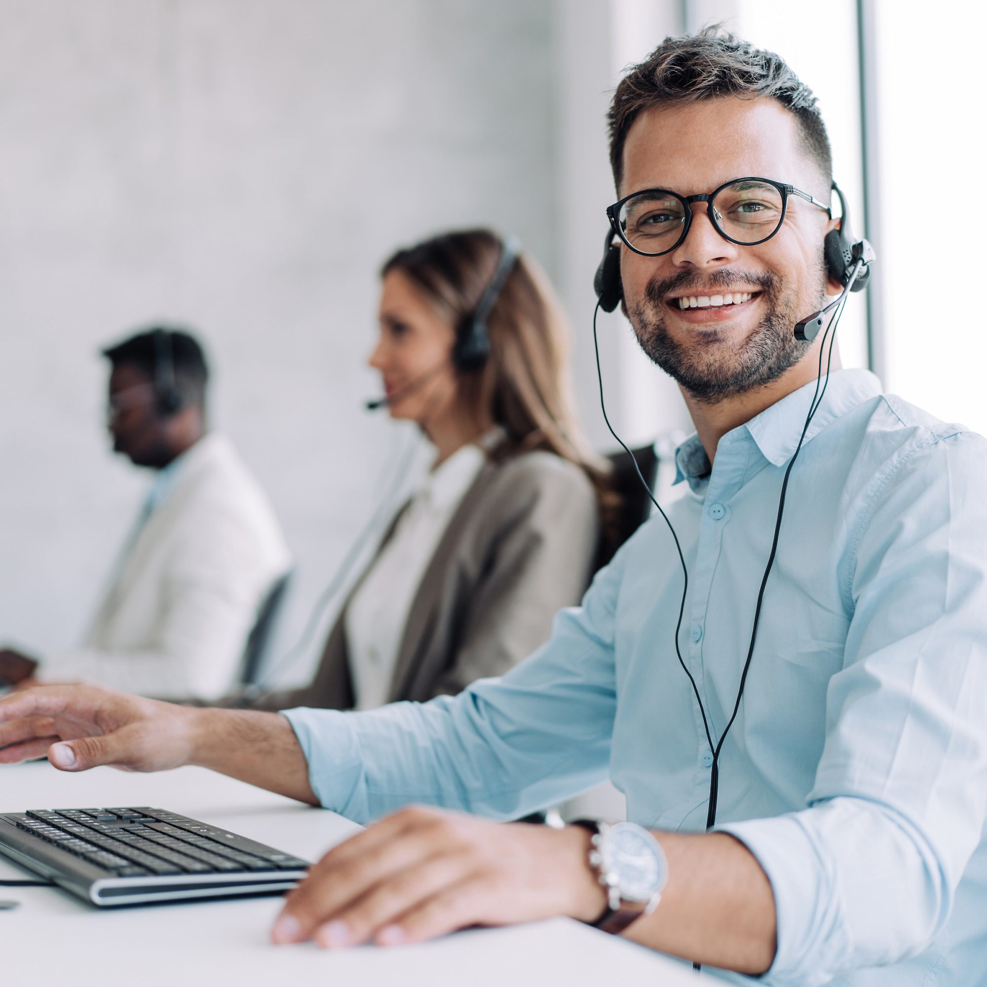 A smiling man with glasses wearing a headset, working at a desk alongside colleagues in a modern office setting.