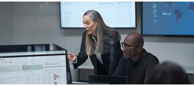 A woman presenting in a meeting with a man and digital screens displaying data in the background.