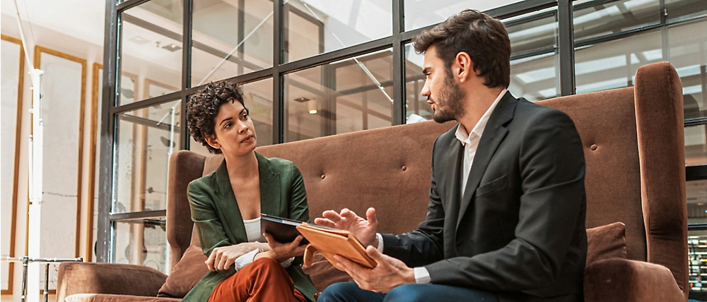 Two professionals discussing over a digital tablet while sitting on a couch in a modern office lobby.