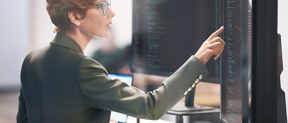 A professional woman in a dark blazer examines and points at data on multiple computer screens in an office setting.