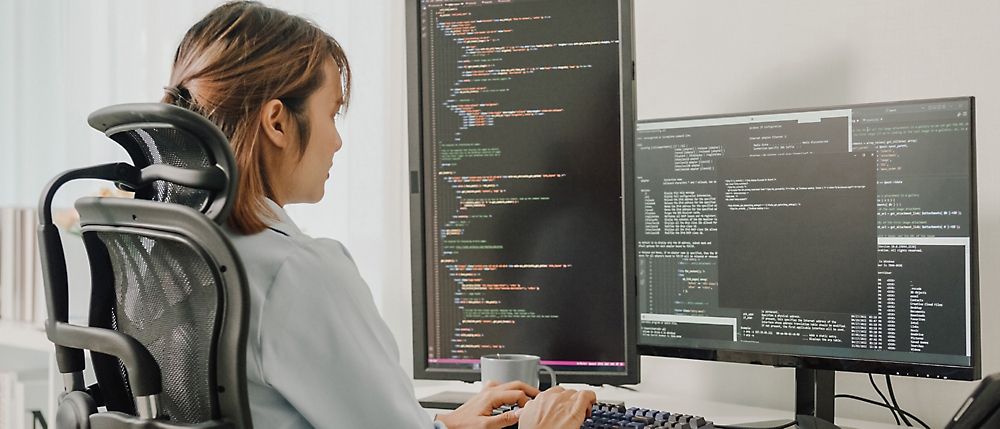 Woman in an office chair reviews code on dual computer screens in a bright workspace.