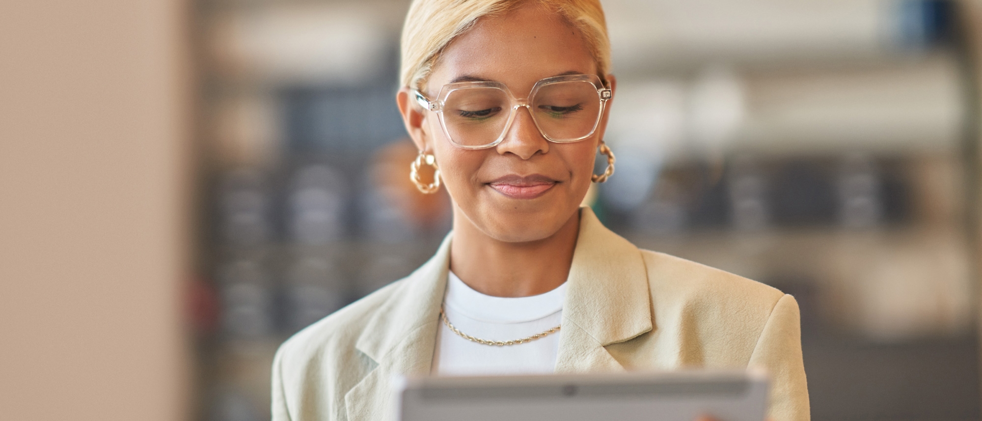 A professional woman with glasses and a light beige blazer reads something on her tablet in a modern office setting.