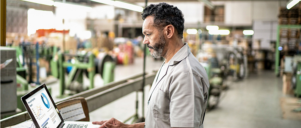 A middle-aged man with gray hair and beard, wearing a light gray shirt, reviews data on a laptop in an industrial warehouse.