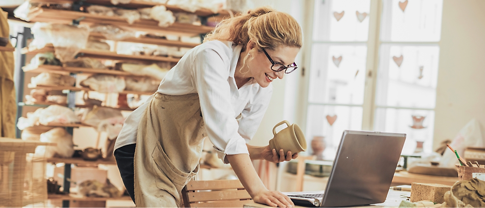 A woman is standing and working on a laptop and holding a cup in her left hand.