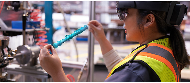 A worker wearing a safety vest and augmented reality glasses inspects an industrial component.