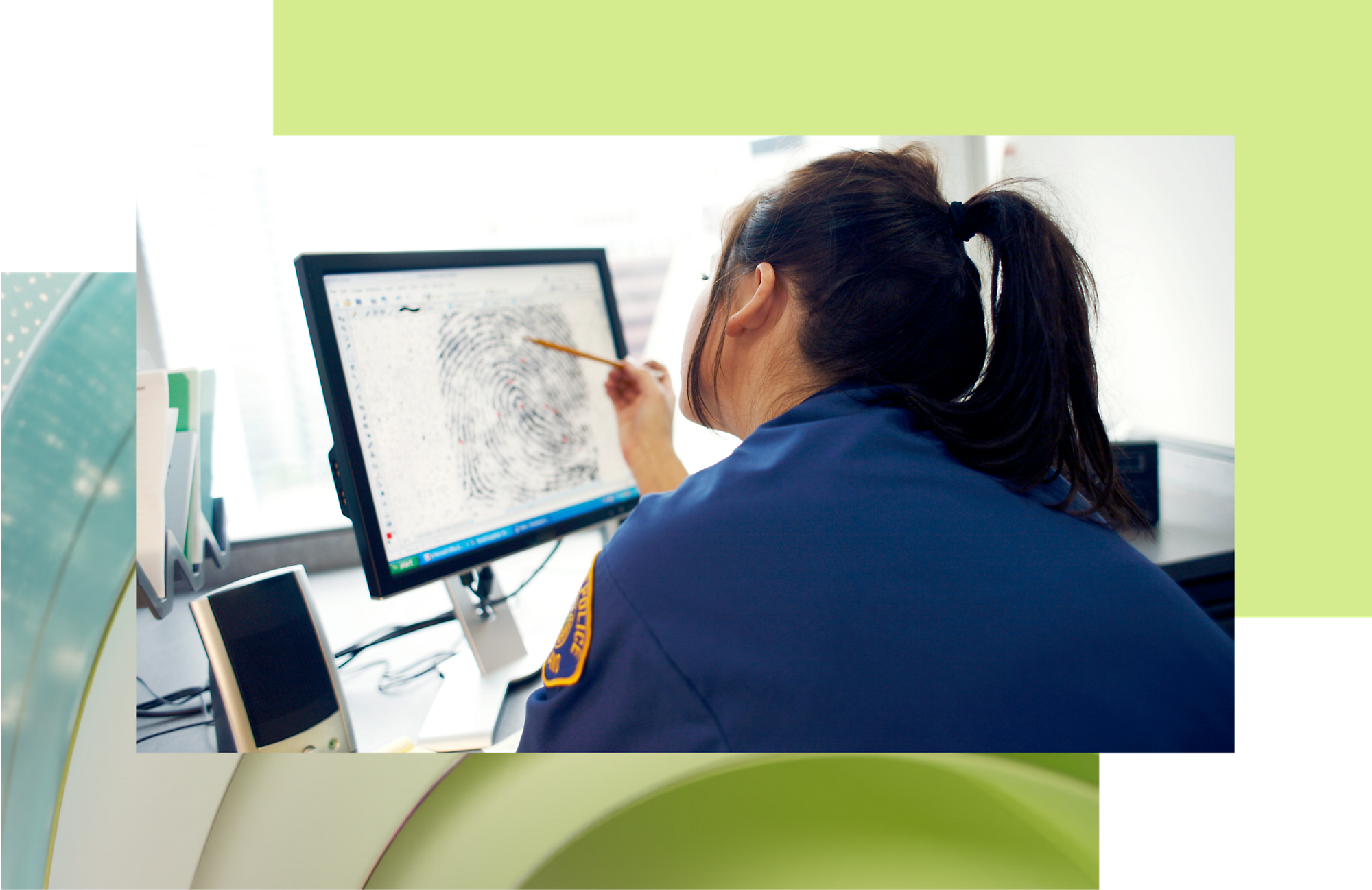 A woman in a blue uniform analyzes a fingerprint on a computer screen in a brightly lit office.