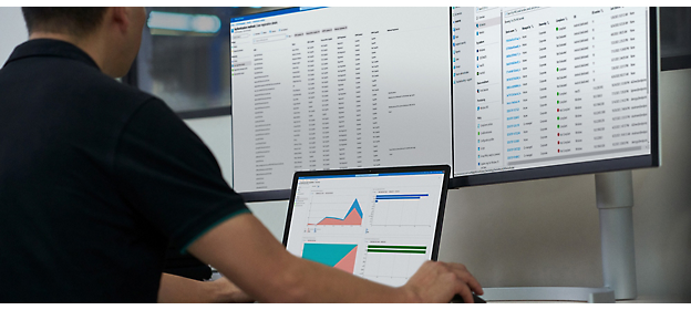 A person working on a laptop with data analysis graphs on the screen, in front of a larger monitor 